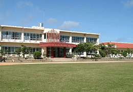 入り口が赤でインパクトのある2階建ての校舎全体を校庭の緑の芝生側から写した写真